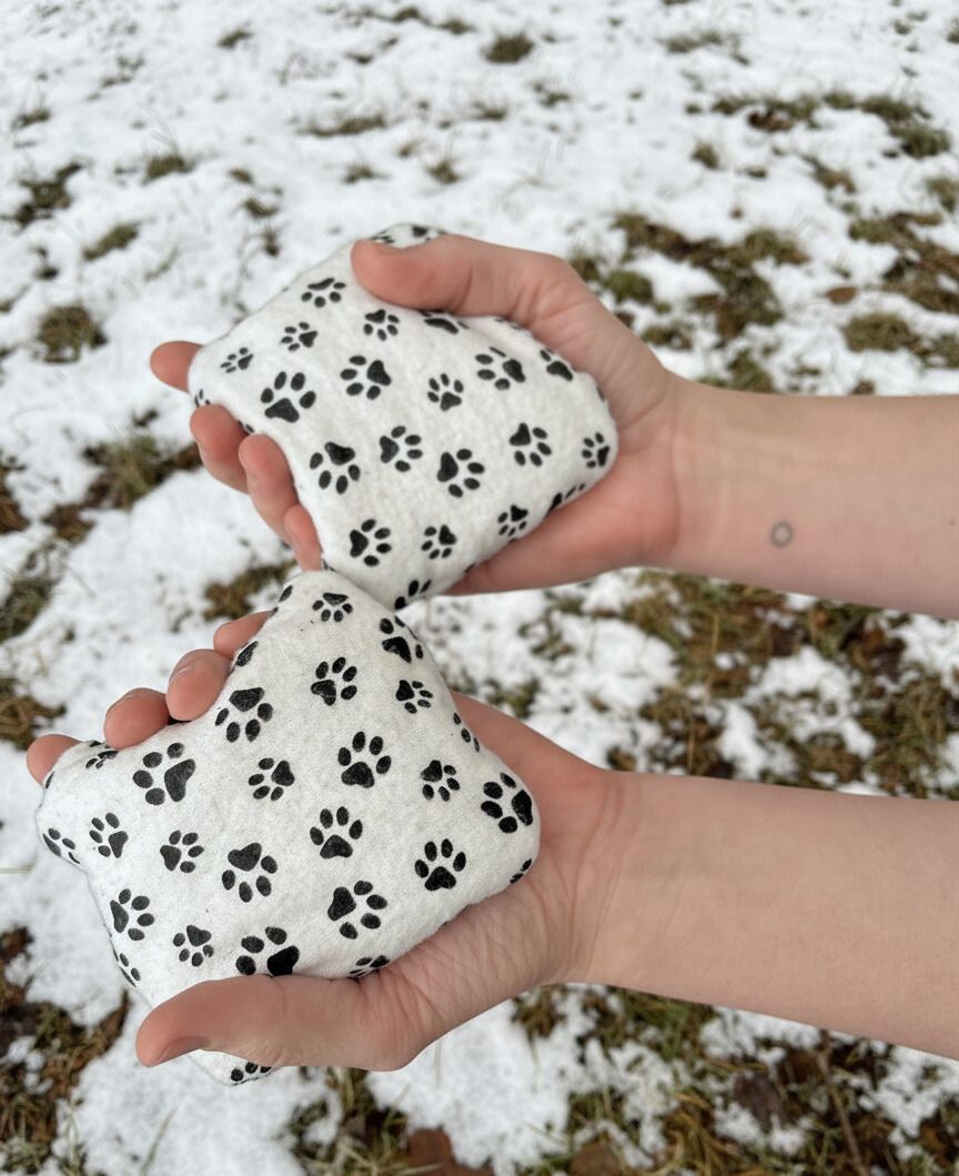 Two white hand warmers with black paw prints held by a person against a snowy ground.